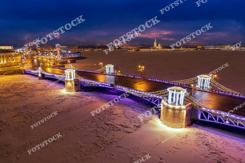 На фотографии изображен Дворцовый мост, украшенный праздничными огнями, которые подчеркивают его архитектурное величие. Заснеженная река и ледяная поверхность контрастируют с ярким освещением моста и окружающих улиц. Этот знаковый мост соединяет центр города с Васильевским островом и является одной из главных достопримечательностей Санкт-Петербурга. Вдалеке видны исторические здания и огни вечернего города, создающие волшебную атмосферу зимнего Петербурга.